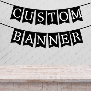 May include: Black banner flags with white text spelling "CUSTOM BANNER" hang against a white wooden background. The banner flags are strung on a black string. A light-colored wooden table is in the foreground.