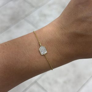 May include: A delicate gold chain bracelet featuring a small, paddle-shaped charm encrusted with tiny, sparkling clear stones. The charm is centered on the thin, polished gold-tone chain, which secures around the wrist with a simple, refined design.
