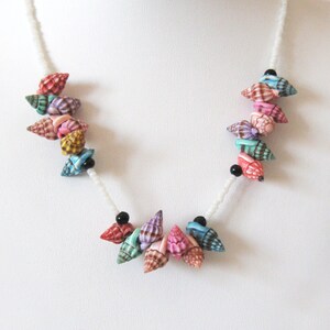 May include: A white beaded necklace with colorful seashells. The shells are arranged in a cluster on either side of the necklace. The shells are a variety of colors, including pink, blue, green, and brown.