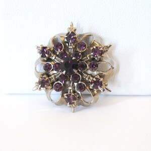 May include: A silver tone brooch with a snowflake design featuring a cluster of purple rhinestones.