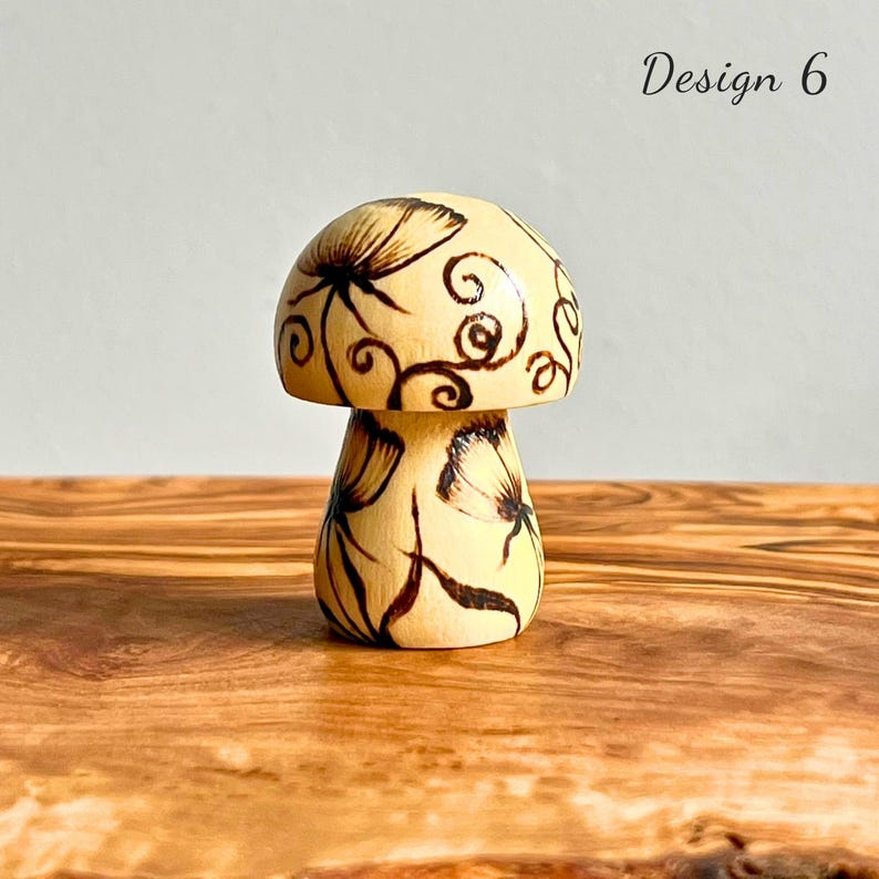 May include: A hand-painted wooden mushroom figurine with a cream-colored cap and stem. The cap features dark brown floral and swirl designs, while the stem has butterfly and floral accents. The words "Design 6" are in the upper right corner.