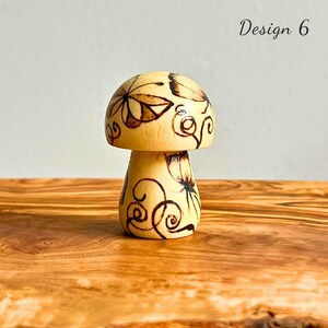 May include: A wooden mushroom figurine with a light brown cap and stem. The cap is decorated with dark brown floral and swirl designs. The text "Design 6" is in the upper right corner. The figurine is on a wooden surface.