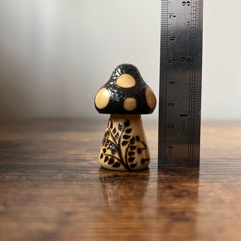 May include: A small, wooden mushroom figurine with a black cap featuring beige spots. The stem is beige with a black leaf design. The ruler next to it shows the mushroom is approximately 2 inches tall.