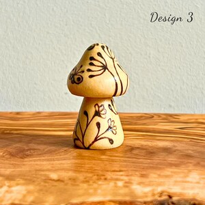 May include: A small, wooden mushroom figurine with a light brown finish. The mushroom cap and stem are decorated with black floral designs. The text "Design 3" is written in black script on the upper right.