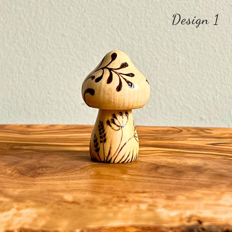 May include: A wooden mushroom figurine with a light brown cap and stem. The cap features dark brown leaf-like designs, while the stem is decorated with floral patterns. The text "Design 1" is visible in the upper right corner.