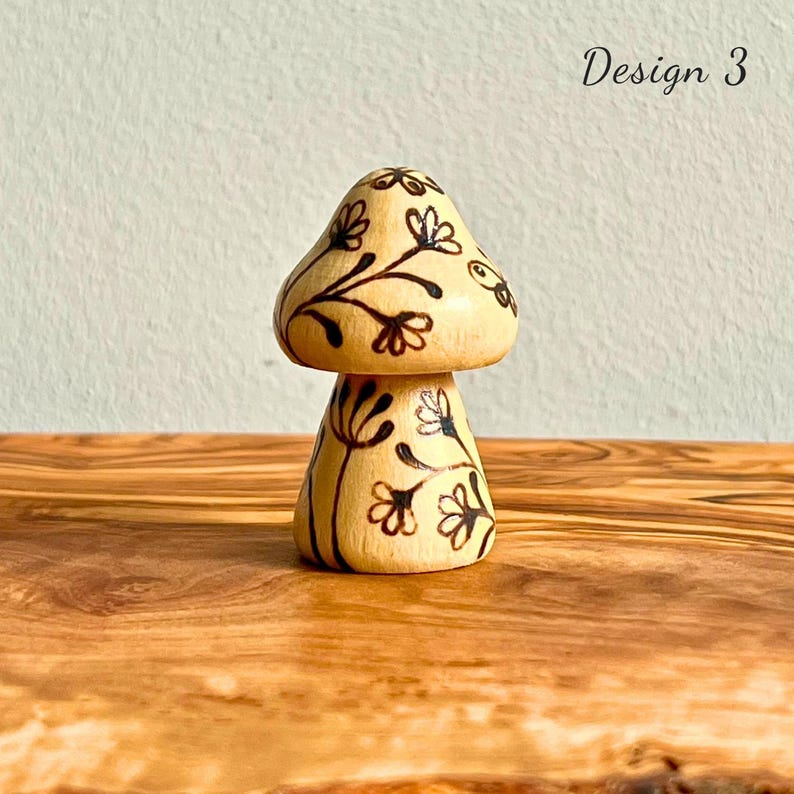 May include: A wooden mushroom figurine with a light brown cap and stem. The cap and stem are decorated with black floral designs. The text "Design 3" is in the upper right corner. The figurine is on a wooden surface.