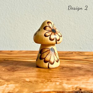 May include: Wooden mushroom figurine with a light brown base and cap. The cap and base are decorated with a brown floral design. The text "Design 2" is written in black on the right side of the image.