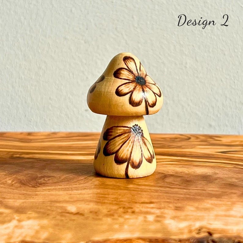 May include: Wooden mushroom figurine with a light brown base and cap. The cap and base are decorated with a brown floral design. The text "Design 2" is written in black on the right side of the image.