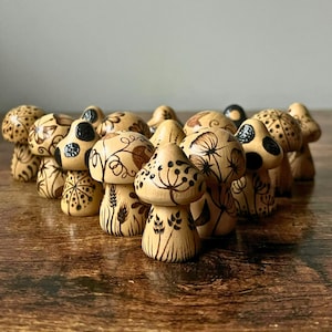 May include: A collection of wooden mushroom figurines with intricate, hand-drawn designs. The mushrooms feature various patterns, including dots, swirls, and floral motifs, in shades of brown and black. The figurines are arranged on a wooden surface.
