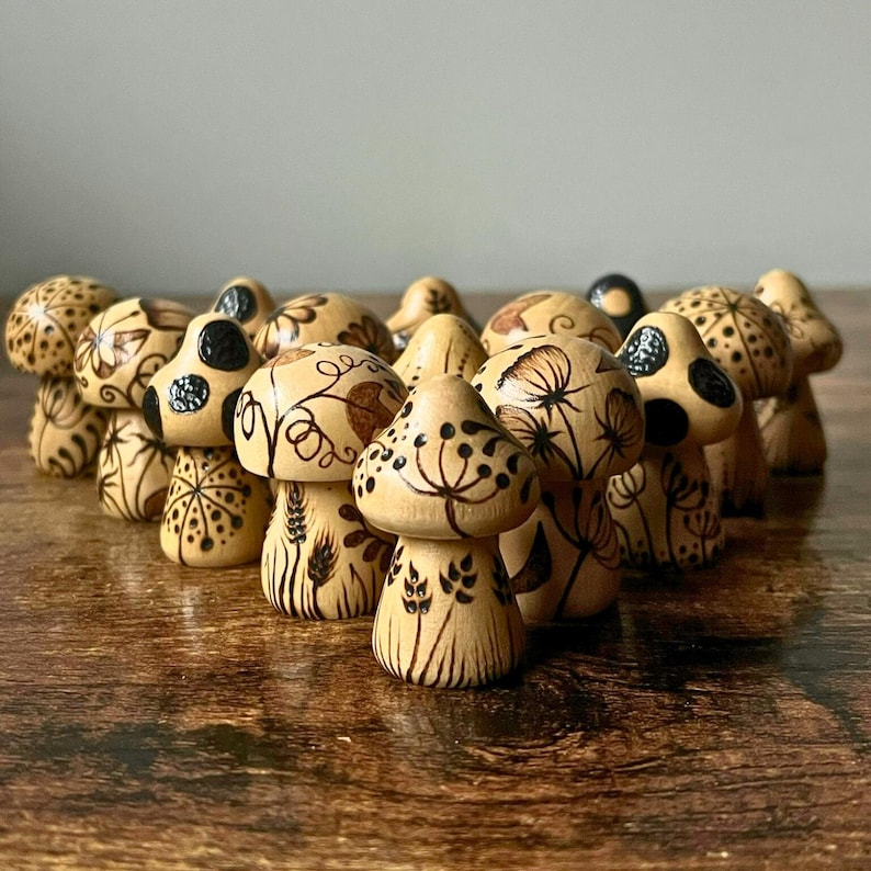 May include: A collection of wooden mushroom figurines with intricate, hand-drawn designs. The mushrooms feature various patterns, including dots, swirls, and floral motifs, in shades of brown and black. The figurines are arranged on a wooden surface.