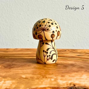 May include: A wooden mushroom figurine with a light brown base and cap. The cap features a dark brown floral design and small black dots. The stem is decorated with black leaves and berries. The text "Design 5" is visible.