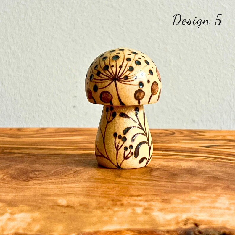 May include: A wooden mushroom figurine with a light brown base and cap. The cap features a dark brown floral design and small black dots. The stem is decorated with black leaves and berries. The text "Design 5" is visible.