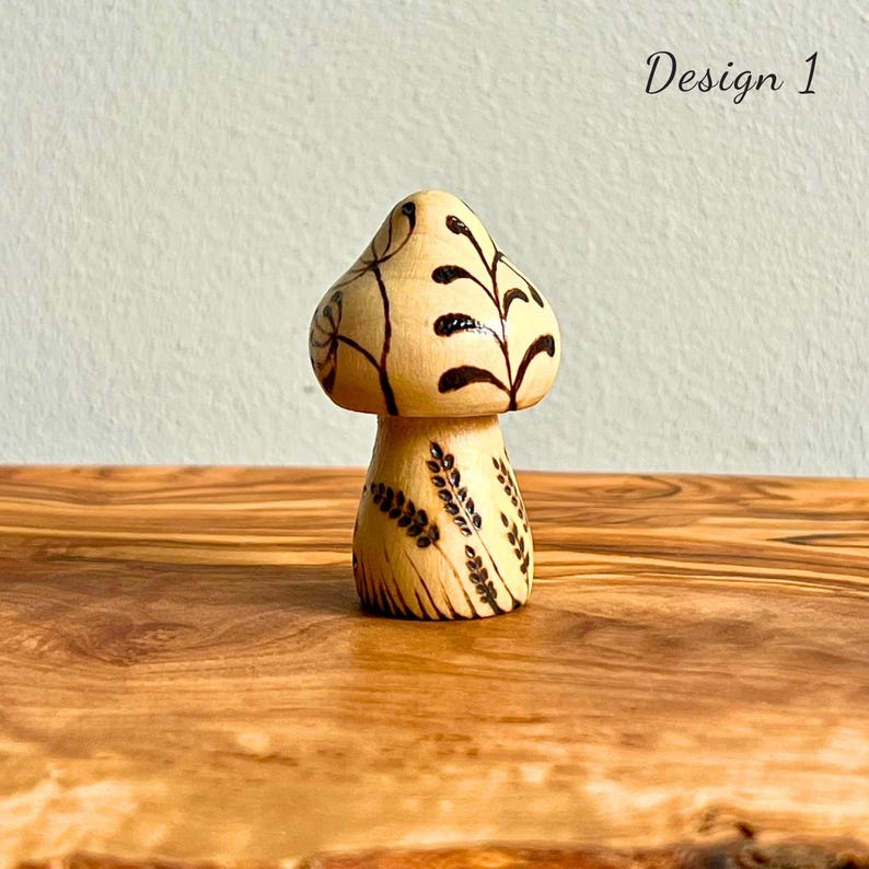 May include: A wooden mushroom figurine with a light brown cap and stem. The cap features dark brown leaf-like designs, while the stem has similar patterns. The text "Design 1" is visible in the upper right corner. The figurine is displayed on a wooden surface.