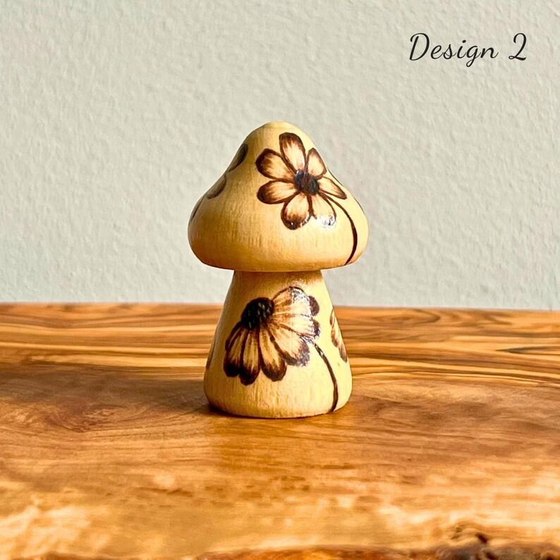 May include: A wooden mushroom figurine with a light brown cap and stem. The cap and stem are decorated with dark brown floral designs. The text "Design 2" is visible in the upper right corner. The figurine is displayed on a wooden surface.