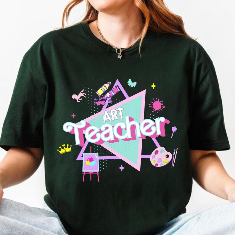 May include: Dark green t-shirt with the words "ART Teacher" in a colorful design. The design includes a pink and teal triangle, a crown, paintbrushes, a palette, and other art-related graphics. The shirt is a casual wear item.