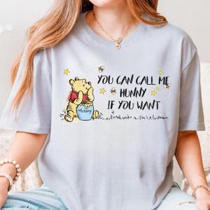May include: Pale blue t-shirt with a graphic of Winnie the Pooh and a honey pot. The text says "YOU CAN CALL ME HUNNY IF YOU WANT" with stars and bees. A casual wear item.