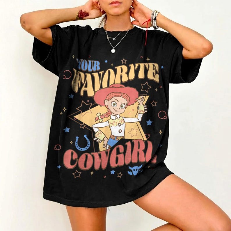 May include: Black t-shirt with a cartoon cowgirl graphic and the text "YOUR FAVORITE COWGIRL." The design includes stars, a horseshoe, and a cowgirl character wearing a pink hat and blue jeans. The shirt has short sleeves.