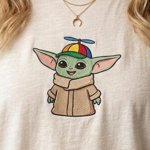 May include: A light beige t-shirt featuring an embroidered graphic of a green creature with large ears wearing a tan robe and a colorful propeller beanie cap with red, yellow, blue, and green panels. A gold-toned crescent moon necklace is visible.