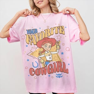 May include: Pale pink t-shirt with a cartoon cowgirl graphic and the words "YOUR FAVORITE COWGIRL". The design incorporates stars, a horseshoe, and a bull's head. The shirt has short sleeves and a round neck.