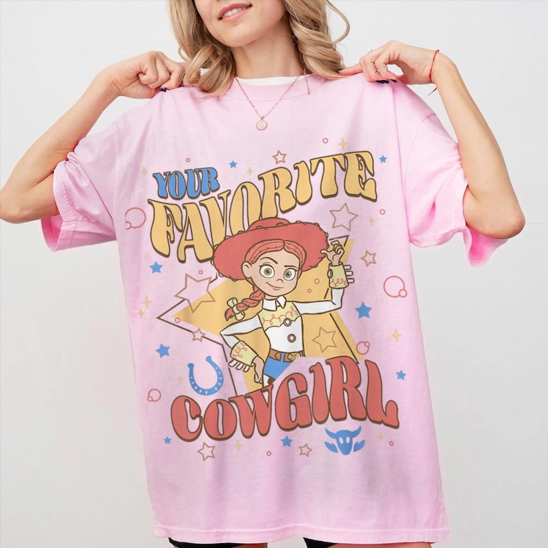 May include: Pale pink t-shirt with a cartoon cowgirl graphic and the words "YOUR FAVORITE COWGIRL". The design incorporates stars, a horseshoe, and a bull's head. The shirt has short sleeves and a round neck.