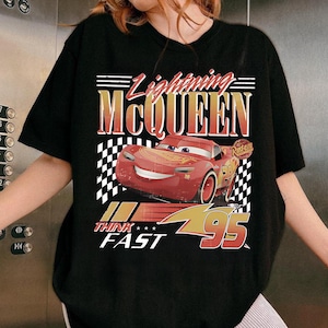 May include: Black t-shirt featuring a graphic of Lightning McQueen from the movie Cars. The design includes the text "Lightning McQueen", "Think Fast", and the number "95" in a racing-themed style. The shirt has a crew neck.