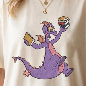 May include: A cream-colored t-shirt featuring a graphic of a purple dragon wearing round glasses. The dragon is smiling and holding a yellow book in one hand and a stack of three books in the other. A small pink flower is near the dragon's tail.