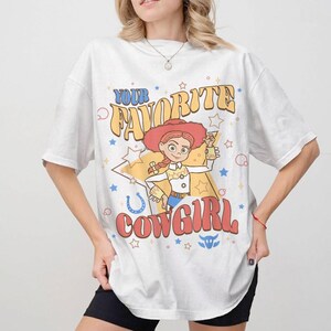 May include: White t-shirt with a retro cartoon cowgirl design and the text "YOUR FAVORITE COWGIRL". The graphic includes stars, a horseshoe, and a bull's head. The cowgirl wears a red hat and holds a small object.