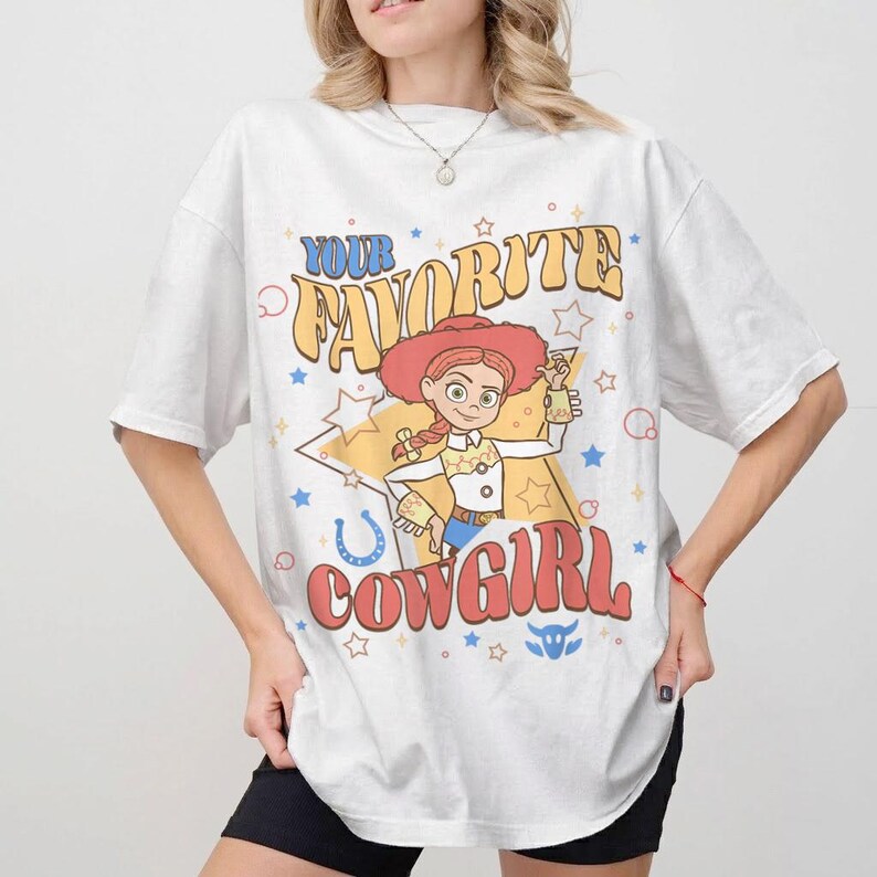May include: White t-shirt with a retro cartoon cowgirl design and the text "YOUR FAVORITE COWGIRL". The graphic includes stars, a horseshoe, and a bull's head. The cowgirl wears a red hat and holds a small object.
