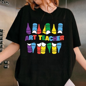 May include: Black t-shirt with a colorful crayon design and the words "ART TEACHER" in rainbow colors. The design features crayon-shaped graphics in various colors, including purple, blue, red, yellow, and green. A fun and creative design for art teachers.