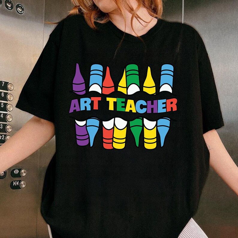 May include: Black t-shirt with a colorful crayon design and the words "ART TEACHER" in rainbow colors. The design features crayon-shaped graphics in various colors, including purple, blue, red, yellow, and green. A fun and creative design for art teachers.