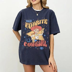 May include: Navy blue t-shirt with a cartoon cowgirl design and the text "Your Favorite Cowgirl". The graphic includes a star, horseshoe, and other decorative elements. The shirt has a relaxed fit.
