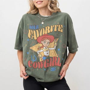 May include: Olive green t-shirt with a graphic of a cowgirl character and the text "YOUR FAVORITE COWGIRL". The design includes stars, a horseshoe, and a star-shaped background. The shirt has short sleeves.
