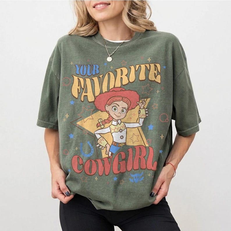 May include: Olive green t-shirt with a graphic of a cowgirl character and the text "YOUR FAVORITE COWGIRL". The design includes stars, a horseshoe, and a star-shaped background. The shirt has short sleeves.
