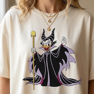 May include: A cream-colored t-shirt featuring an embroidered graphic of a duck character dressed in a black and purple robe with large horns, holding a gold staff. The character has a white face, orange beak, and is wearing a high-collared cape.