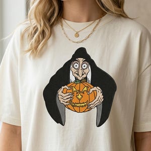 May include: An off-white t-shirt featuring a large embroidered graphic of a witch with pale skin, a black hooded cloak, and long fingers holding an orange jack-o'-lantern with a carved face. The design is centered on the chest of the garment.