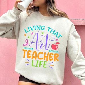 May include: A light gray sweatshirt with the text "LIVING THAT Art TEACHER LIFE" in various colors. The design includes stars, pencils, and an apple. The sweatshirt is a casual, comfortable garment.