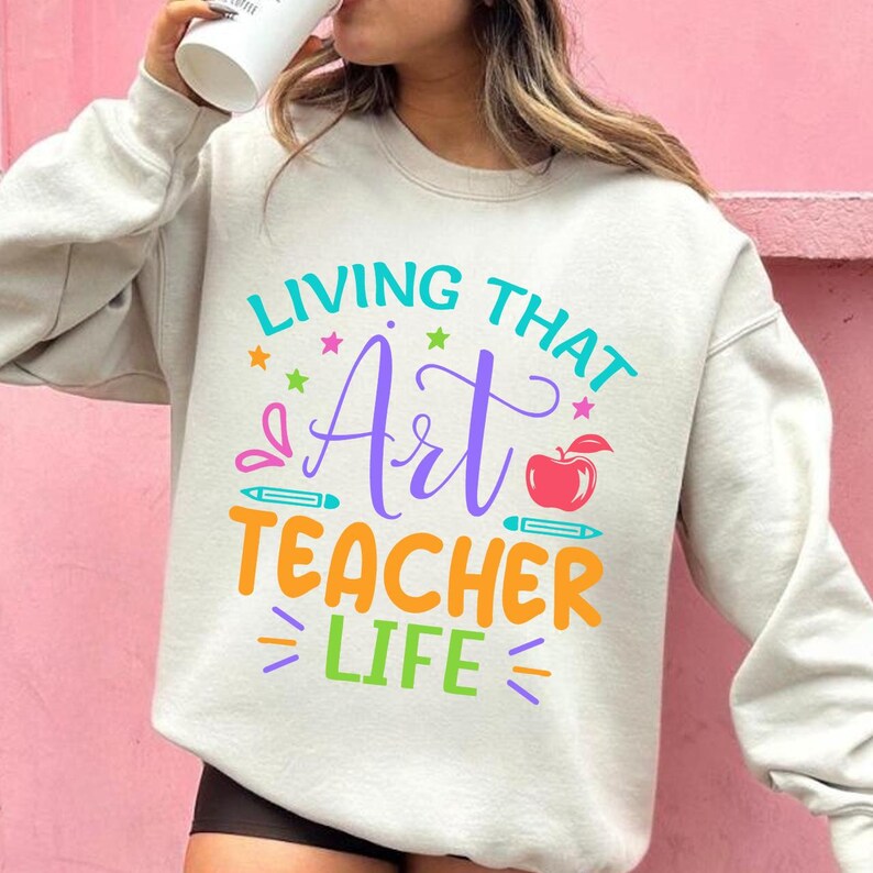 May include: A light gray sweatshirt with the text "LIVING THAT Art TEACHER LIFE" in various colors. The design includes stars, pencils, and an apple. The sweatshirt is a casual, comfortable garment.