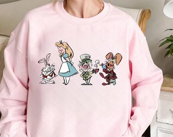May include: A light pink sweatshirt featuring a colorful graphic of characters from Alice in Wonderland. The graphic includes Alice, the White Rabbit, the Mad Hatter, and the March Hare. The sweatshirt is made of a soft material.