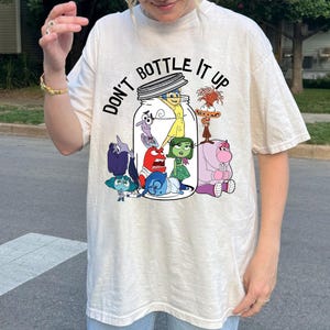 May include: Cream-colored t-shirt featuring the phrase "DON'T BOTTLE IT UP" above a cartoon illustration of characters inside a jar. The characters are from the movie Inside Out. The shirt has short sleeves and a relaxed fit.