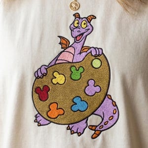 May include: A cream-colored t-shirt featuring an embroidered purple dragon character holding a large brown artist palette. The palette is decorated with six colorful, mouse-shaped paint splatters in red, orange, yellow, green, blue, and purple.
