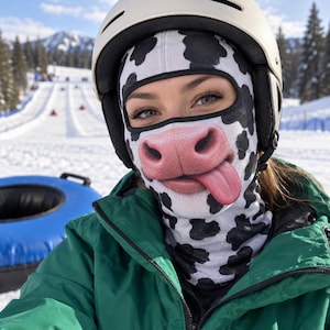 May include: A balaclava featuring a black and white cow print pattern with a realistic graphic of a pink cow nose and a tongue sticking out. The fabric covers the head and neck, leaving an opening for the eyes, worn under a white ski helmet.