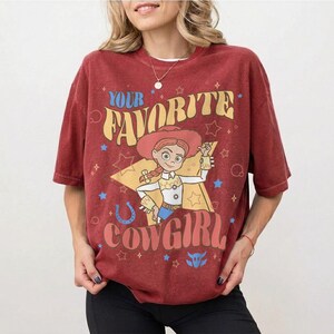 May include: A maroon t-shirt with a graphic of a cowgirl character and the text "YOUR FAVORITE COWGIRL" in a retro font. The design includes stars, a horseshoe, and a bull icon. The shirt has short sleeves and a crew neck.