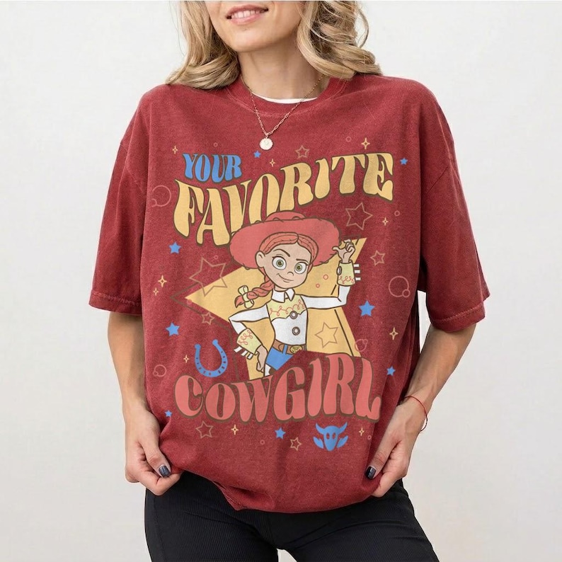 May include: A maroon t-shirt with a graphic of a cowgirl character and the text "YOUR FAVORITE COWGIRL" in a retro font. The design includes stars, a horseshoe, and a bull icon. The shirt has short sleeves and a crew neck.