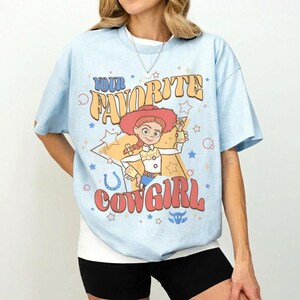 May include: Pale blue t-shirt with a cartoon cowgirl graphic and the text "YOUR FAVORITE COWGIRL". The design includes stars, a horseshoe, and a cowgirl character in a red hat. The shirt is a light blue colour.