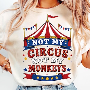 May include: Cream-colored t-shirt featuring a circus tent design in red and white stripes, with the text "NOT MY CIRCUS NOT MY MONKEYS" in a playful font. The design includes stars and banners.