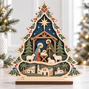 Könnte beinhalten: Eine mehrschichtige Weihnachtsbaum-Dekoration aus Holz mit einer Krippenszene, die Maria, Josef und das Jesuskind in einer Krippe zeigt. Das Design umfasst drei weiße Schafe, Tannenbäume, einen Stern und eine Wüstenstadt mit Palmen.