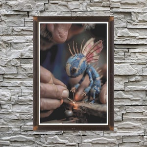 May include: A framed image of a blue and tan creature with pink and blue wings being worked on with a welding torch. The creature is in the center of the image, with the hands of a person working on it.