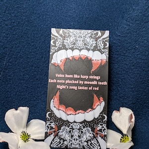 May include: A black card with a vampire teeth design and the text "Veins hum like harp strings, Each note plucked by moonlit teeth, Night's song tastes of red." Two white flowers with yellow centers are on the left and right.