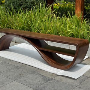 May include: A dark brown wooden bench featuring a continuous, curved base that forms an integrated lower shelf. The bench has a smooth, polished finish and a long, flat seat surface that extends horizontally across the curved support structure.