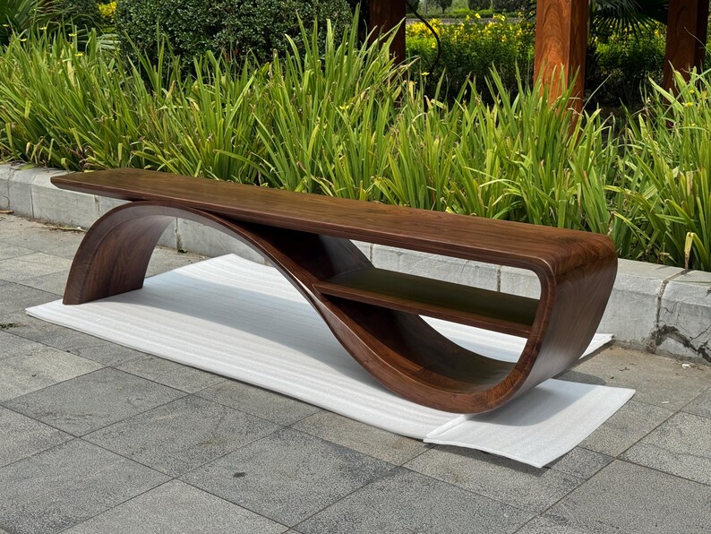 May include: A dark brown wooden bench featuring a continuous, curved base that forms an integrated lower shelf. The bench has a smooth, polished finish and a long, flat seat surface that extends horizontally across the curved support structure.